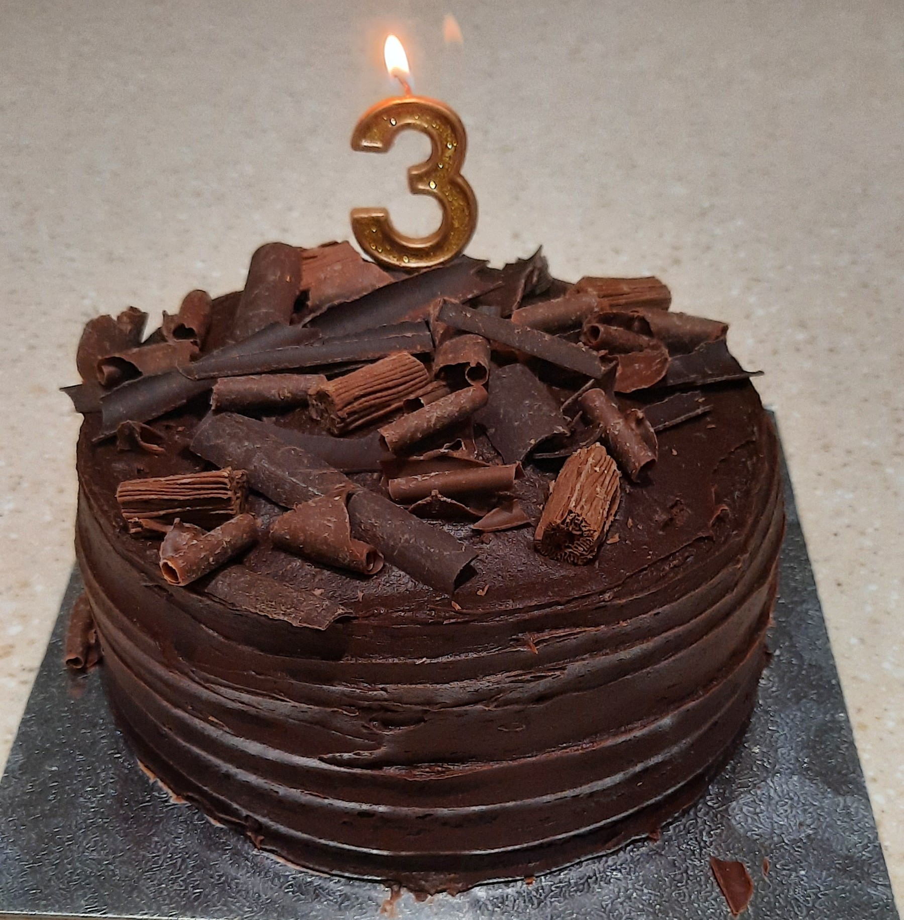 A round chocolate cake with a three shaped candle burning.